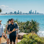 A group of people walk along a coastal path with city skyscrapers in the background and the ocean to their right on a sunny day.