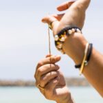 Close-up of a person’s hands tying a thin string, with colorful beaded bracelets on their wrist, against a blurred outdoor background with water and sky.