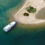 A blue and white boat is docked at a small sandy island surrounded by greenish-blue water, viewed from above.