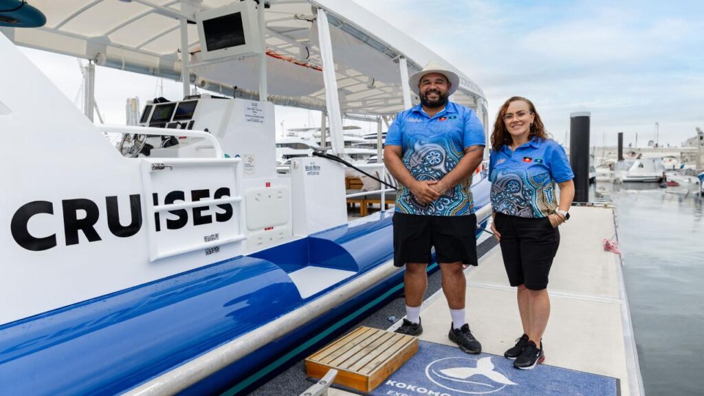 Two people in matching blue uniforms stand on a dock next to a white cruise boat with the word "CRUISES" partially visible on the side.