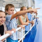 A group of children stand on a boat deck, looking and pointing out at the water, with the sea and sky visible in the background.