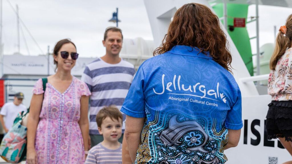 A woman wearing a "Jellurgal Aboriginal Cultural Centre" shirt stands facing a family of three near a docked boat.