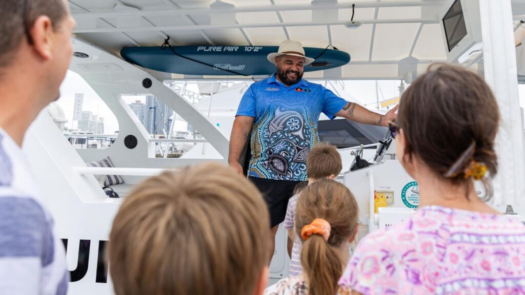 A man stands on a boat talking to a small group of people, including children, with a city skyline and docked boats visible in the background.