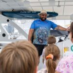 A man stands on a boat talking to a small group of people, including children, with a city skyline and docked boats visible in the background.