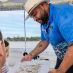 A man in a hat and patterned shirt shows a map to two people on a boat, with water and trees visible in the background.