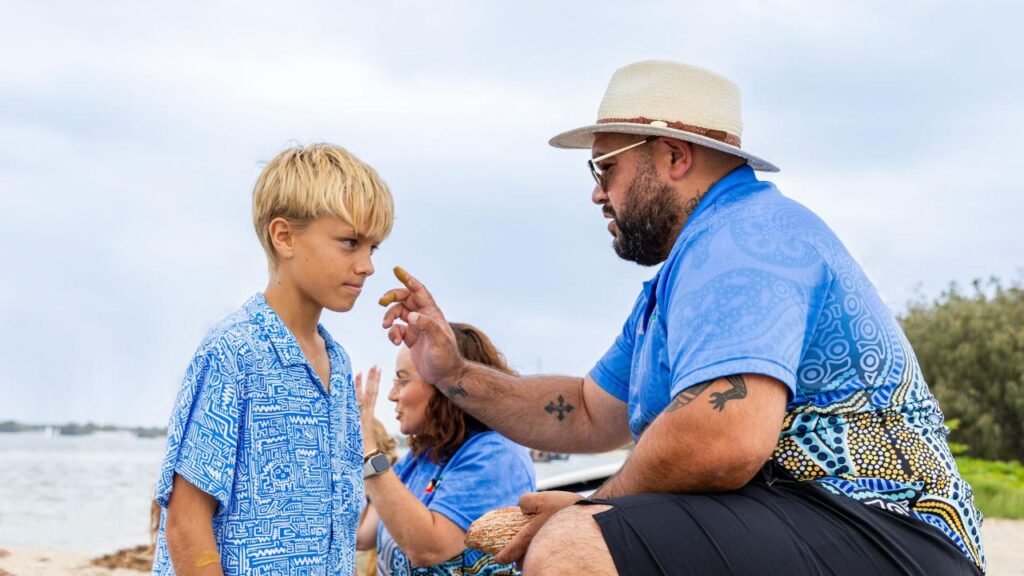 An adult in a patterned blue shirt applies ochre to a boy’s forehead near a beach, while another person sits in the background.