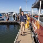 A man and a woman walk on a dock beside a red boat, with other people and boats visible in the marina under a partly cloudy sky.