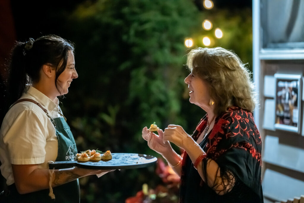 A woman in an apron holds a tray of appetizers while another woman talks to her and holds a bite-sized snack, outdoors at night.