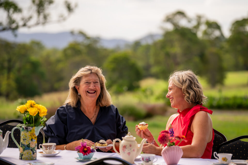 Two women sit at an outdoor table decorated with flowers and tea sets, smiling and enjoying pastries with a green, blurred landscape in the background.
