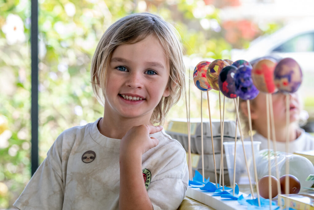 A young child with shoulder-length blonde hair smiles beside a display of decorated lollipops on sticks at an outdoor event.