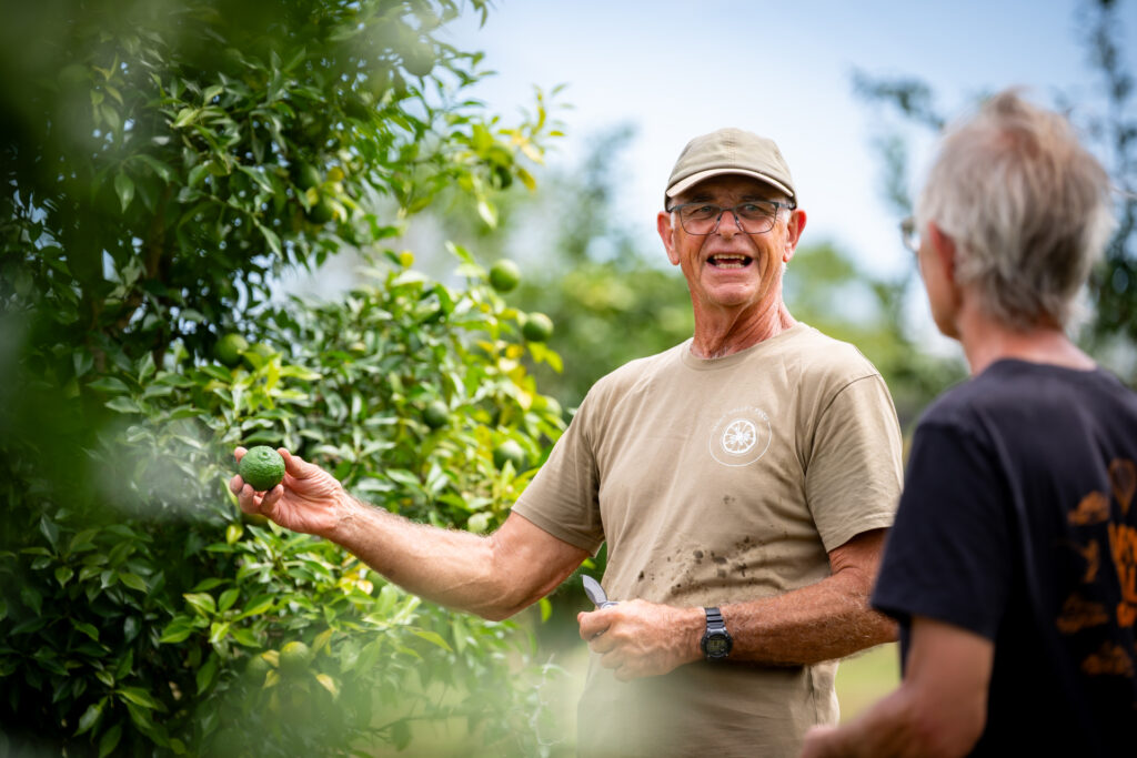 Two men stand near a leafy tree; one man holds a green fruit and smiles while talking to the other.