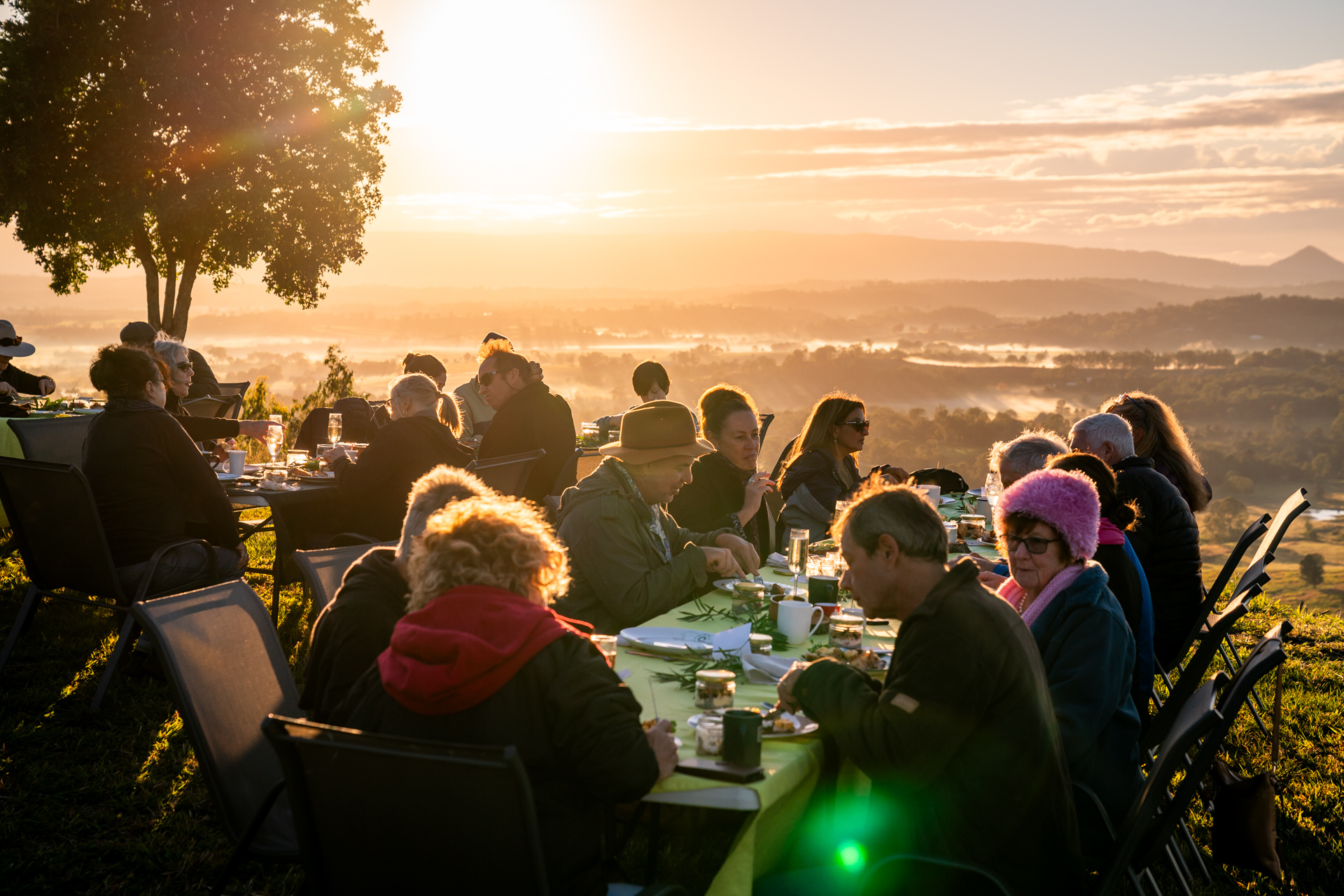 A group of people sit at outdoor tables eating breakfast together at sunrise, with scenic hills and trees in the background.