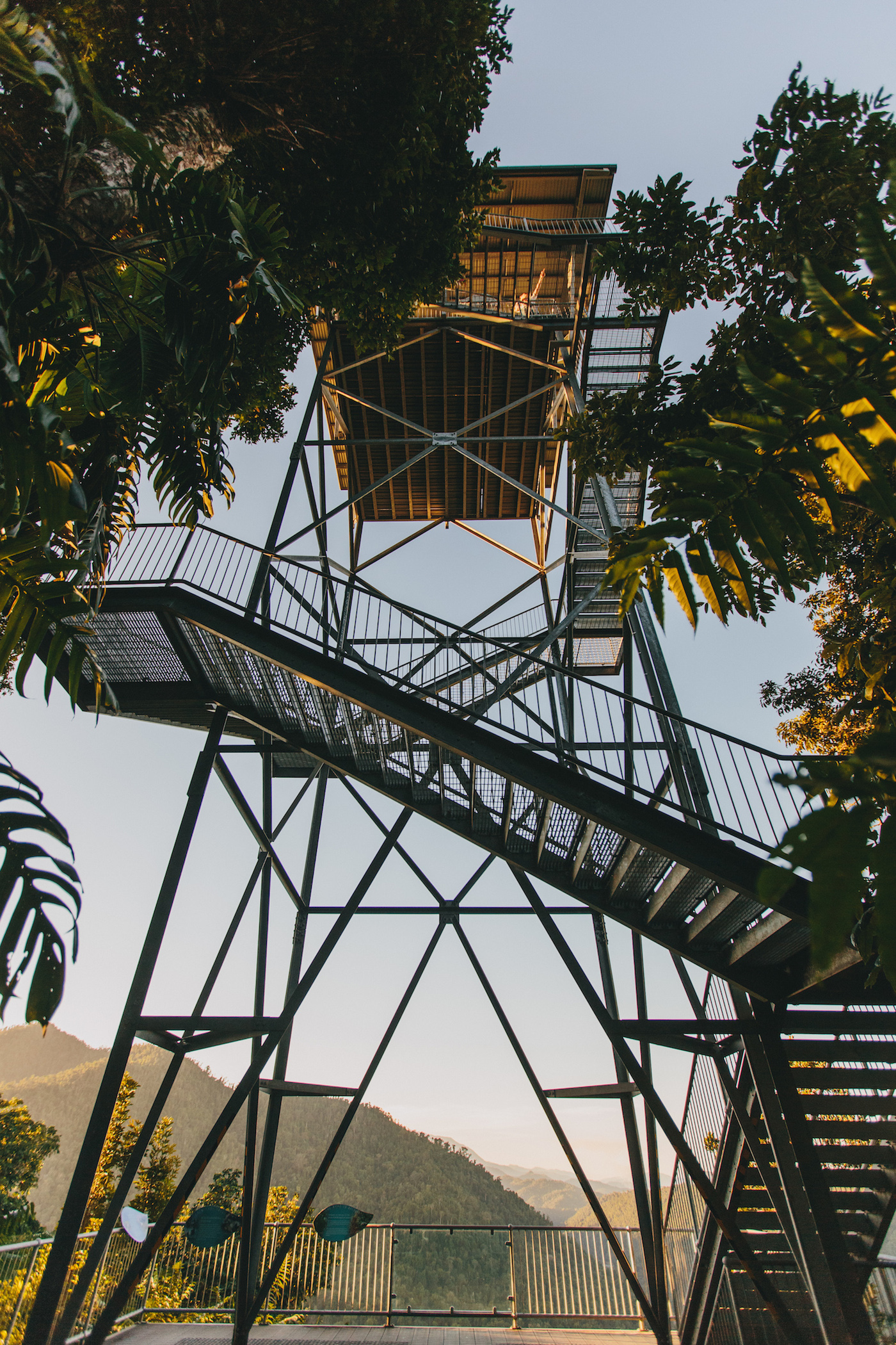 A tall metal observation tower with stairs, surrounded by trees, stands against a clear sky with mountains visible in the background.