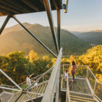 Three people stand on a metal observation platform overlooking a forested valley and distant mountains during sunset.