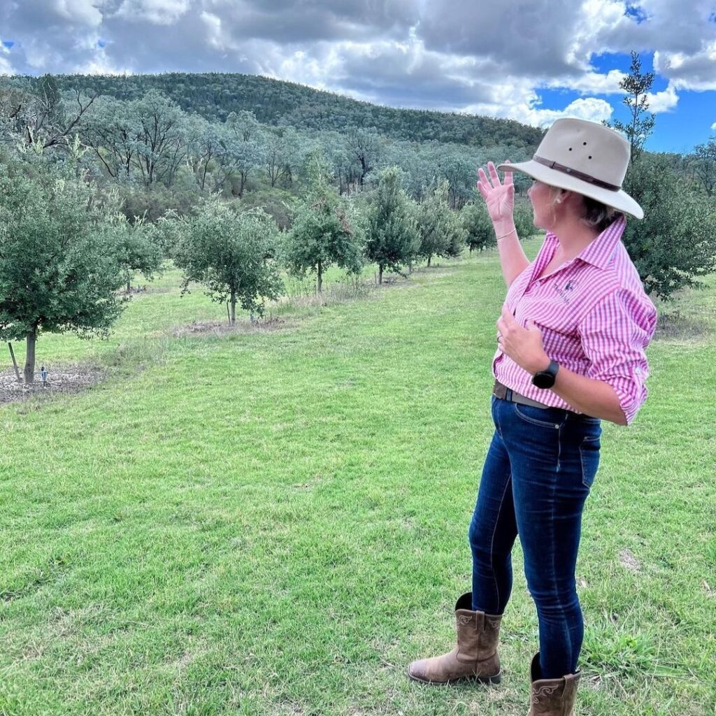 A woman in a hat, pink shirt, jeans, and boots stands on grass, gesturing toward a green, tree-covered hill under a partly cloudy sky.