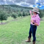 A woman in a hat, pink shirt, jeans, and boots stands on grass, gesturing toward a green, tree-covered hill under a partly cloudy sky.
