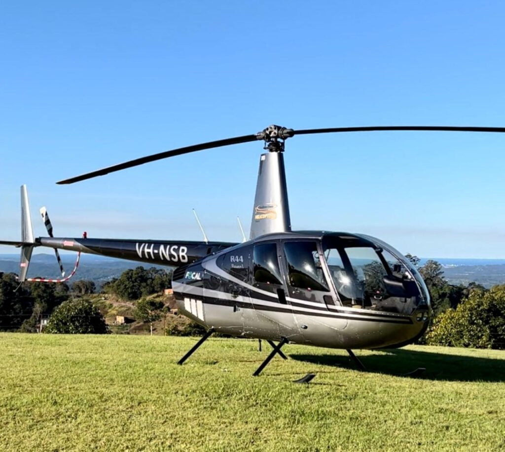 A silver and black Robinson R44 helicopter with registration VH-NSB is parked on a grassy field under a clear blue sky.