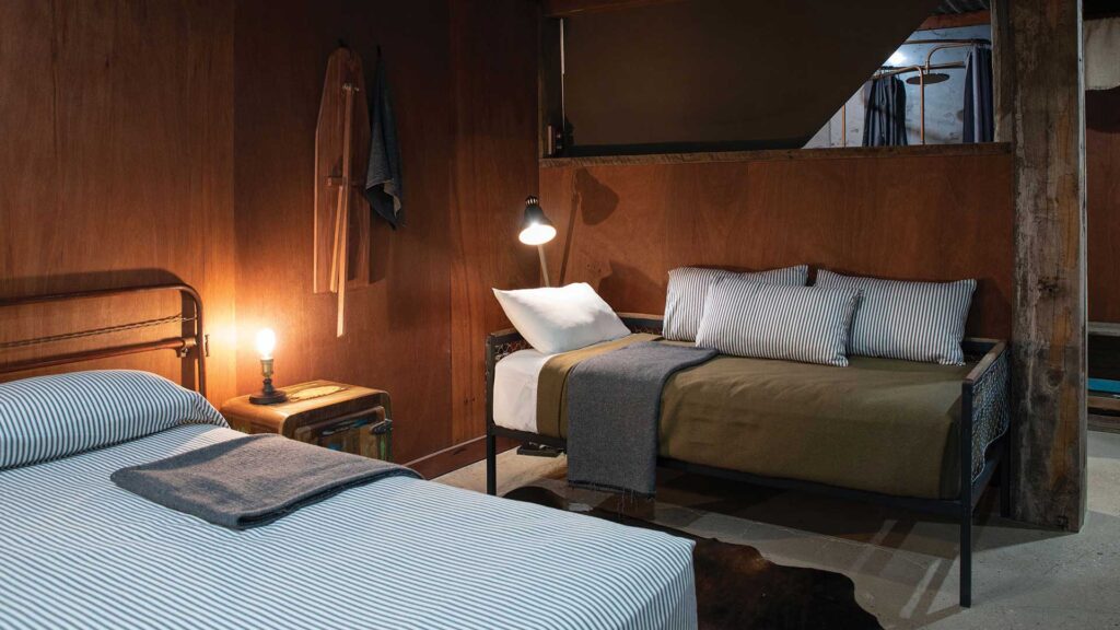 A cozy bedroom with two single beds, each with striped bedding, a small nightstand with a lamp, and wooden walls. A robe and clothes hang on the wall in the background.