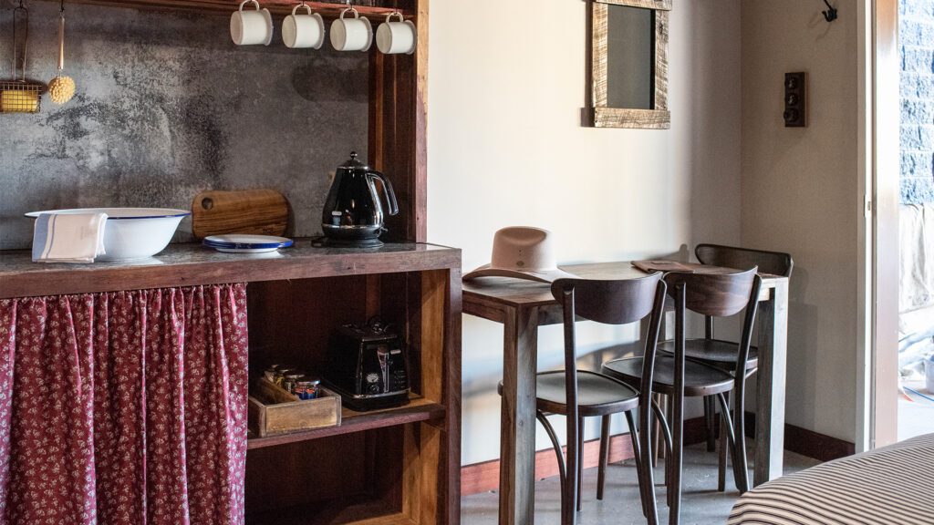 A small kitchen and dining area with wooden furniture, a kettle, cups hanging above the counter, and two chairs at a table next to a wall with a mirror.