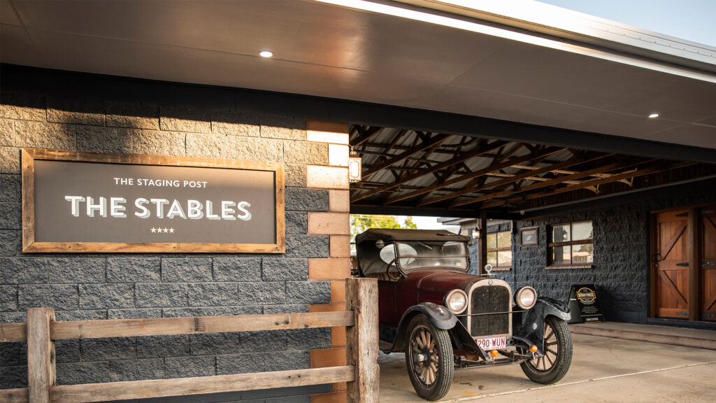 An old-fashioned car is parked under a wooden roof next to a building with a sign that reads 