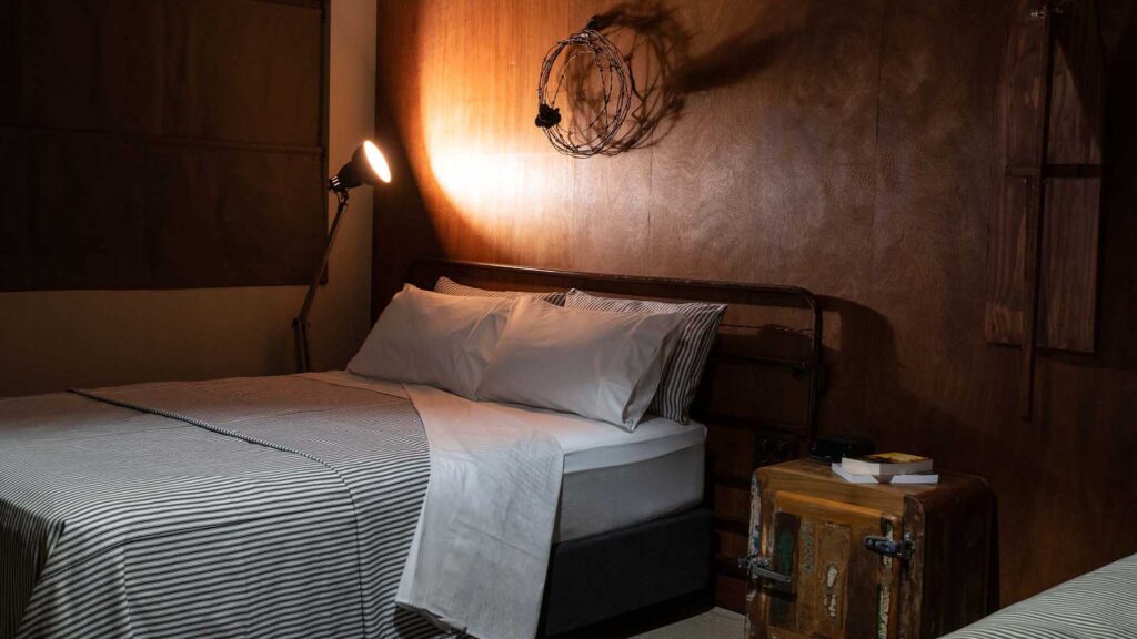 A neatly made bed with striped bedding is beside a vintage trunk used as a nightstand, a lamp, and wall art in a dimly lit, modern rustic bedroom.