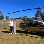 A man and a woman stand next to a black R44 Raven II helicopter on a grassy field with a vineyard and clear blue sky in the background.