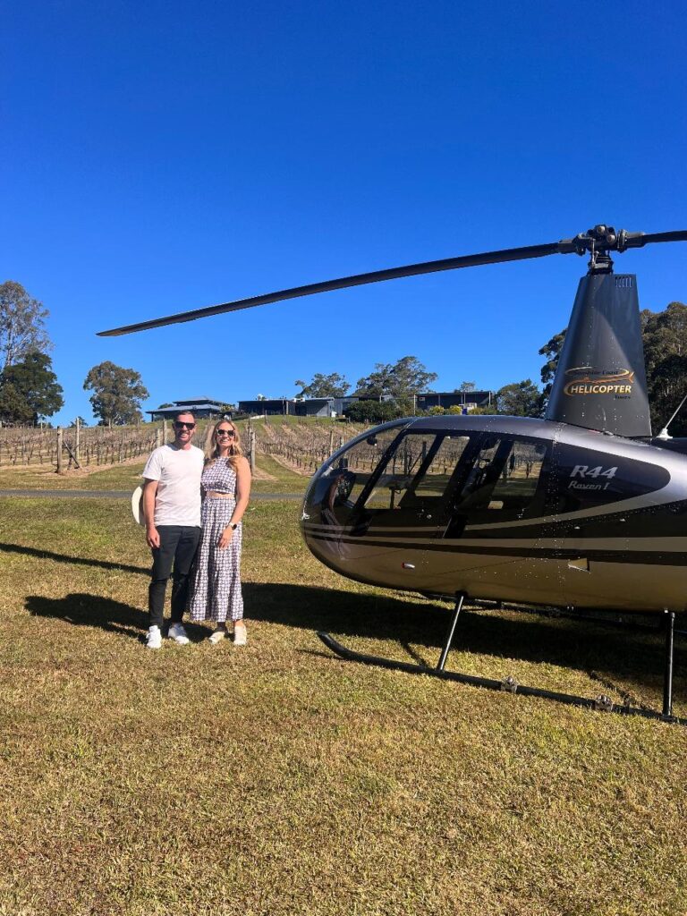 A man and a woman stand next to a black R44 Raven II helicopter on a grassy field with a vineyard and clear blue sky in the background.