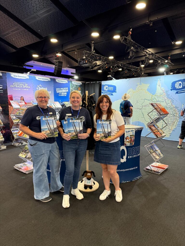 Three people stand indoors at a tourism booth, each holding brochures in front of a display rack and a large map backdrop. A small stuffed dog sits on a stand in front of them.
