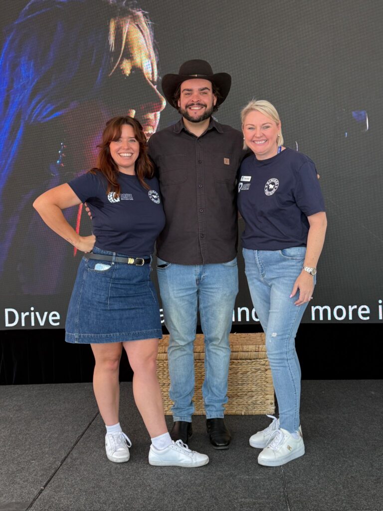 Three people stand on a stage smiling, with two women in matching blue shirts and denim skirts or shorts, and a man in a cowboy hat and black shirt, against a screen background.