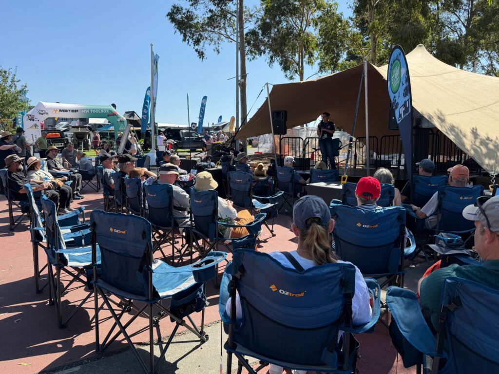 A group of people sits in camping chairs facing a stage where a person speaks into a microphone under a tent at an outdoor event.
