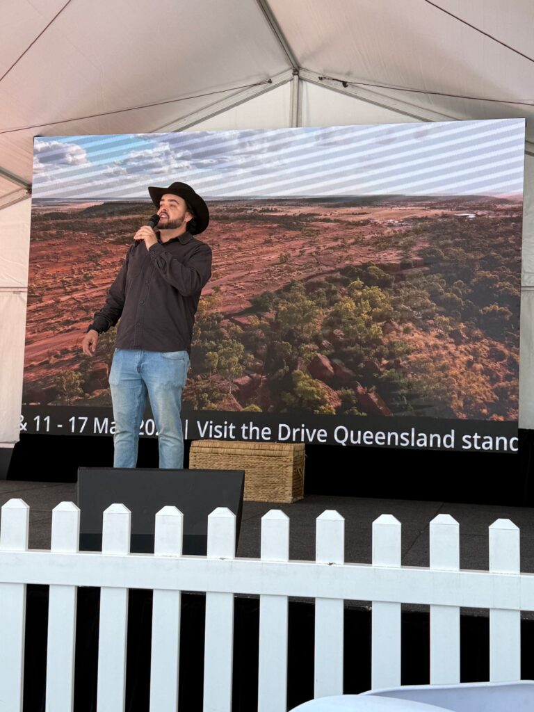 A man in a black hat and jacket speaks on stage with a microphone, standing in front of a large screen displaying a landscape image.