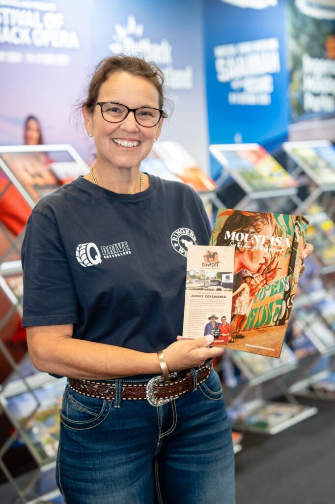 A woman wearing glasses and a navy t-shirt stands indoors, smiling and holding two brochures in front of a display of pamphlets and posters.