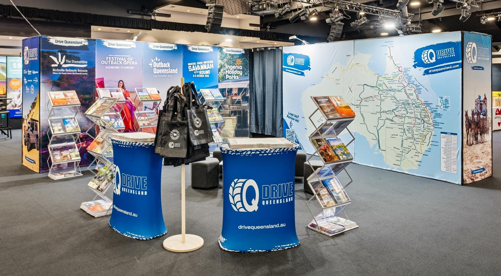 Exhibition booth for Drive Queensland featuring brochures, maps, display stands, promotional bags, and a large map of Queensland on the wall.
