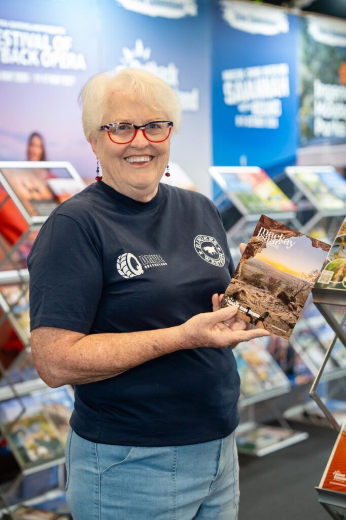 An older woman with short white hair and glasses holds a travel magazine while standing in front of a display of brochures.