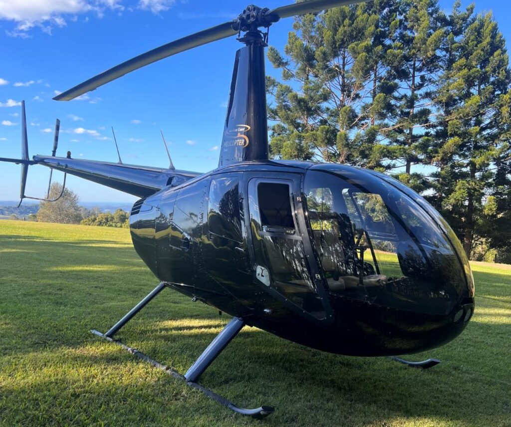 A black helicopter is parked on a grassy field with trees and a blue sky in the background.