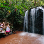 A family sits near a small waterfall in a lush, green forest, with three children observing the water and parents seated beside them.