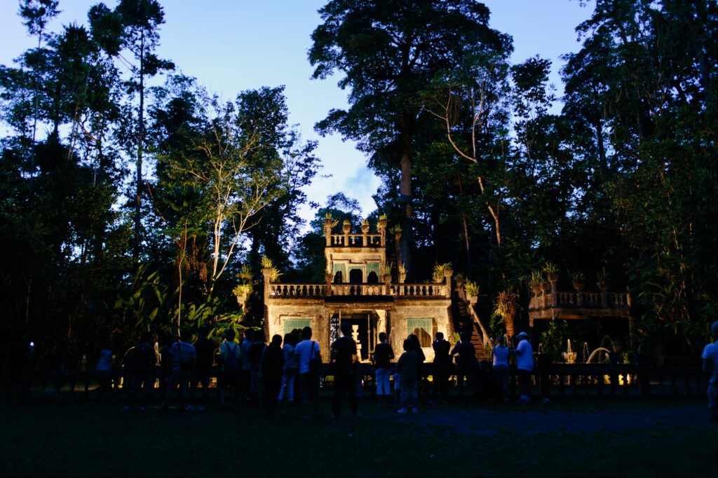A group of people stands in front of an ornate, castle-like structure surrounded by dense trees at dusk.