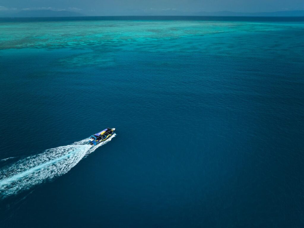 A speedboat creates a white wake as it travels across deep blue water toward a lighter turquoise reef in the distance under a clear sky.