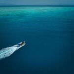 A speedboat creates a white wake as it travels across deep blue water toward a lighter turquoise reef in the distance under a clear sky.