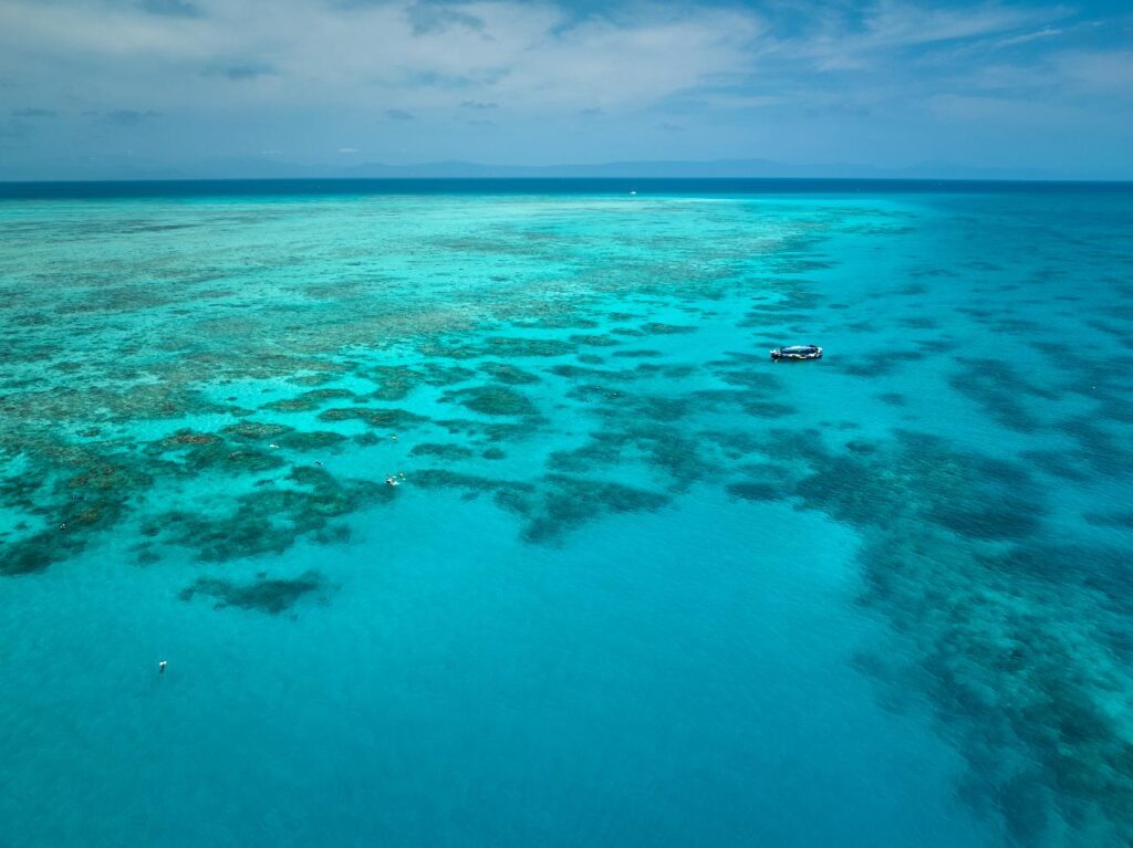 Aerial view of a boat floating on clear turquoise water above coral reefs, with small patches of darker coral visible beneath the surface.