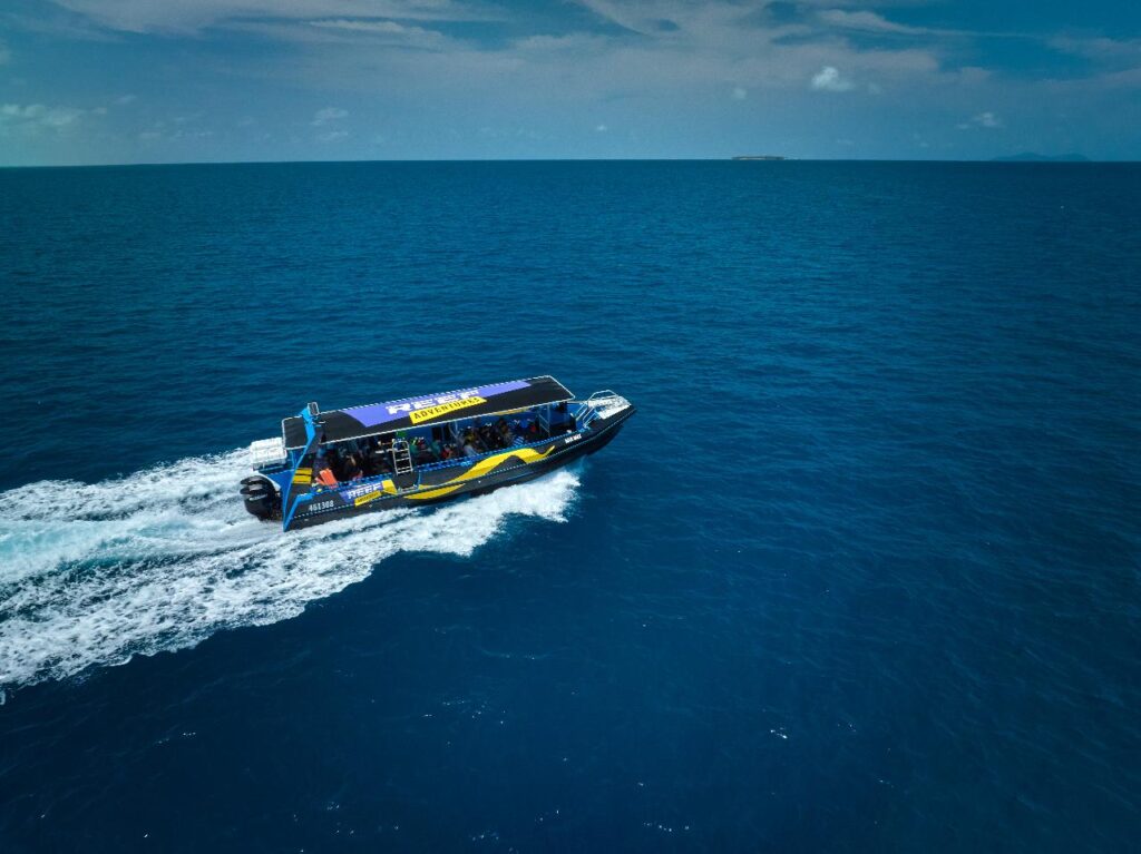 A passenger speedboat moves across calm blue ocean water under a partly cloudy sky, leaving a white wake behind.