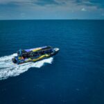 A passenger speedboat moves across calm blue ocean water under a partly cloudy sky, leaving a white wake behind.