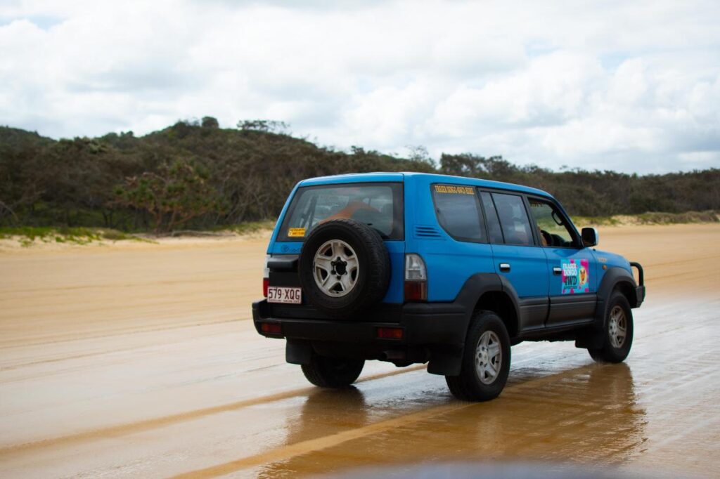 A blue SUV with stickers is driving on a sandy beach with forested hills in the background under a cloudy sky.