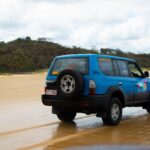 A blue SUV with stickers is driving on a sandy beach with forested hills in the background under a cloudy sky.