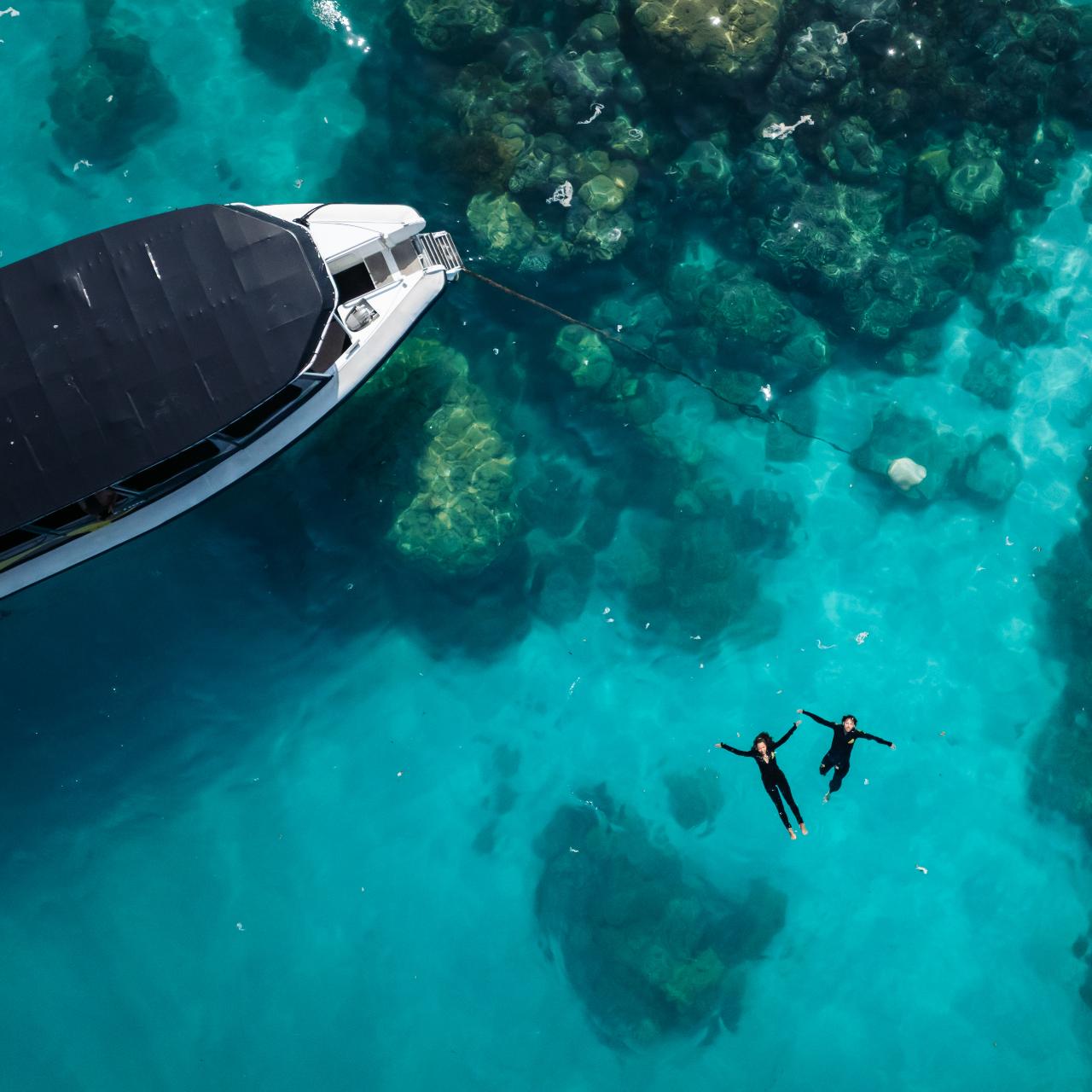 Aerial view of two people snorkeling near a boat in clear blue water with visible underwater rocks.