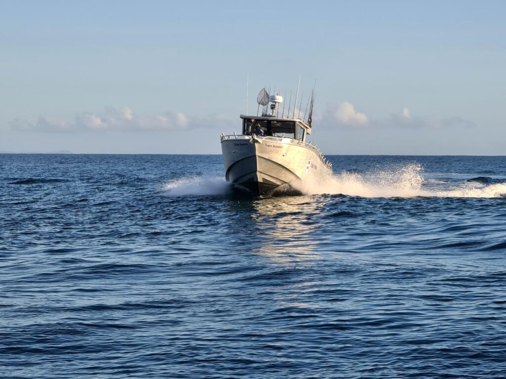 A white motorboat moves quickly across blue open water, creating waves and white spray behind it under a clear sky.