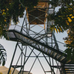 Tall metal observation tower with staircases, surrounded by trees, and overlooking a distant mountain landscape under a clear sky.