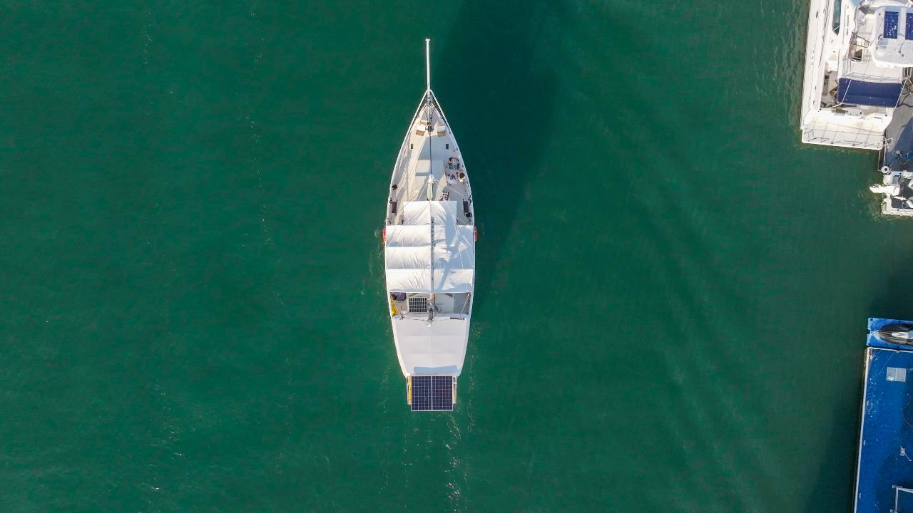 Aerial view of a white sailboat with solar panels floating on calm green water, docked near other boats.