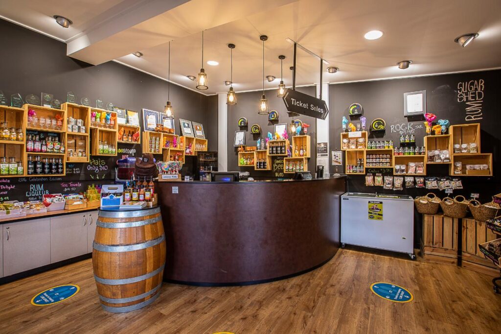 A shop interior with a curved counter, shelves displaying various food products, a wooden barrel, hanging lights, and a sign reading “Ticket Sales.”.