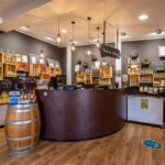 A shop interior with a curved counter, shelves displaying various food products, a wooden barrel, hanging lights, and a sign reading “Ticket Sales.”.